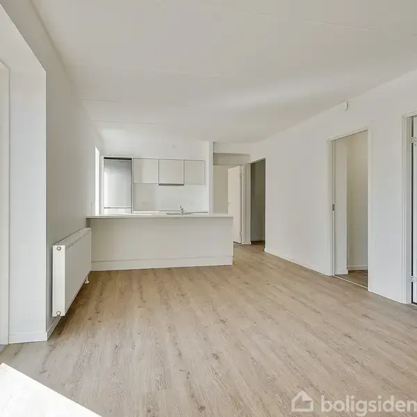 Et lyst rum med trægulv og hvide vægge indeholder en åben køkkenafdeling. Store vinduer på venstre side giver naturligt lys. En åben dør fører til et andet rum.