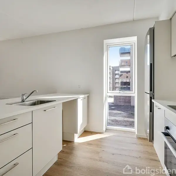 Et lyst køkken med hvide skabe og skuffer, indbygget ovn og kogeplade til højre. En stor vindue bader rummet i sollys, og der er udsigt til en bygning udenfor.