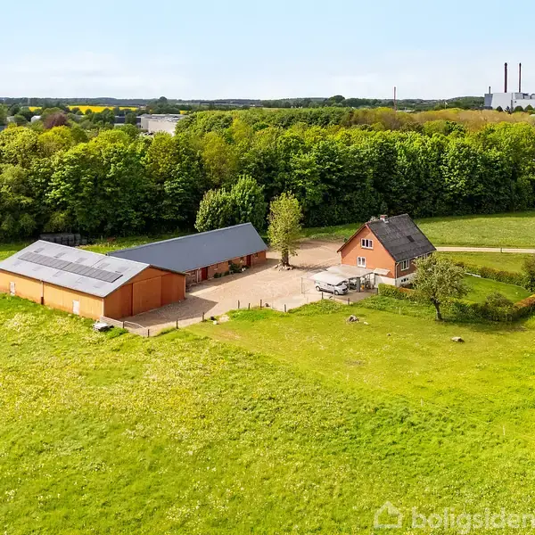 To bygninger ligger på en stor græsplæne omgivet af træer; en moderne lade og et rødt murstenshus med sort tag. I baggrunden ses nogle industrielle strukturer.