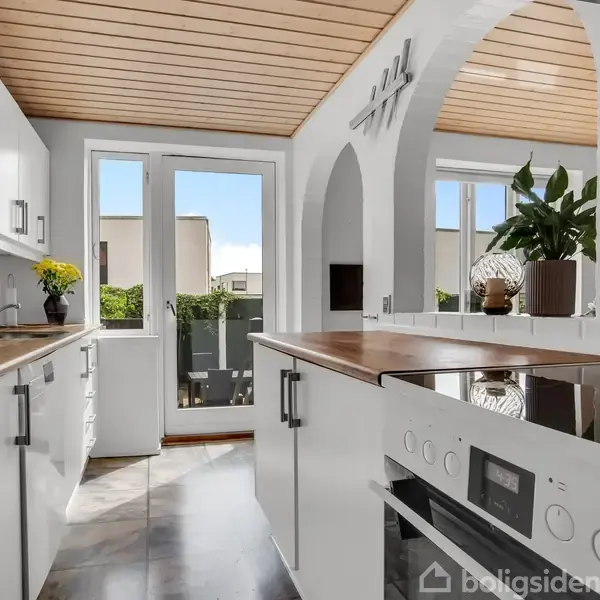 Et hvidt, moderne køkken med træloft og indbyggede skabe. Store vinduer giver udsigt til en terrasse og have. Plante dekorationer pryder rummet, der er lyst og luftigt.