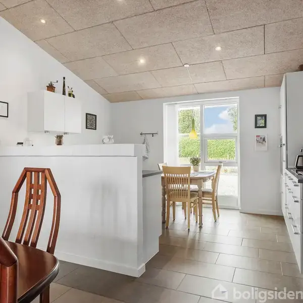 Et køkkenalrum med spisebord og stole ved et vindue. Køkkenskabe og apparater er i højre side, mens en væg adskiller spise- og køkkenområderne. Vægge og lofter er lyse.