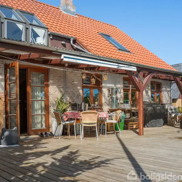 Terrassen har borde og stole under en træoverdækning, mens planter og grill tilføjer hygge. Murstenshuset med rødt tag og ovenlysvinduer danner baggrunden med solrige omgivelser.