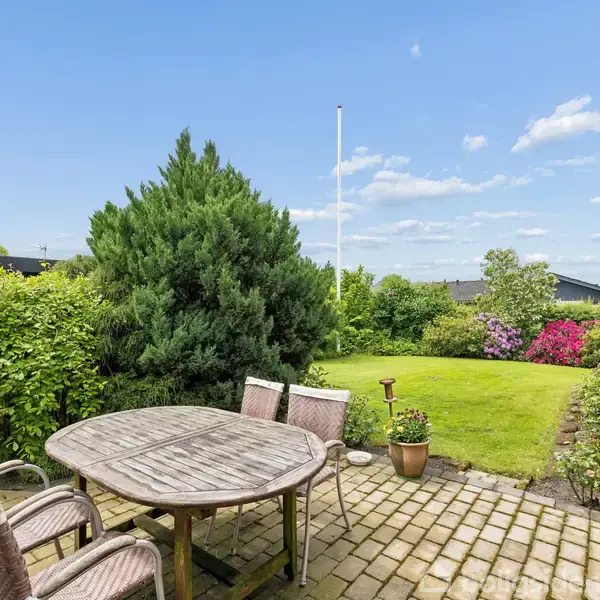 Et rundt bord med stole står på en terrasse af brudsten. En velplejet have med græsplæne og blomsterbede omgiver terrassen, og en flagstang ses i baggrunden under en blå himmel.