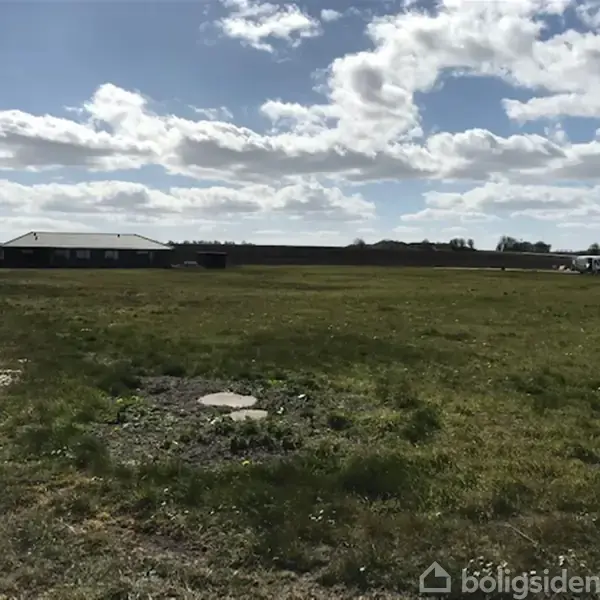 Græsområde under en blå himmel med hvide skyer, en enkelt lygtepæl til venstre, huse og en bil i baggrunden.