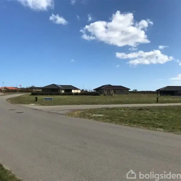 Et asfalteret vejforløb drejer gennem en åben forstadskontekst med lavtliggende huse omkring. Græsarealer kantet af vejen og en blå himmel med spredte skyer. Ingen tekst til stede.