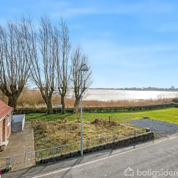 Et rødt murstenshus med solpaneler på taget står stille ved en vej, tæt på en græsplæne og træer. I baggrunden ses en sø under en klar himmel.
