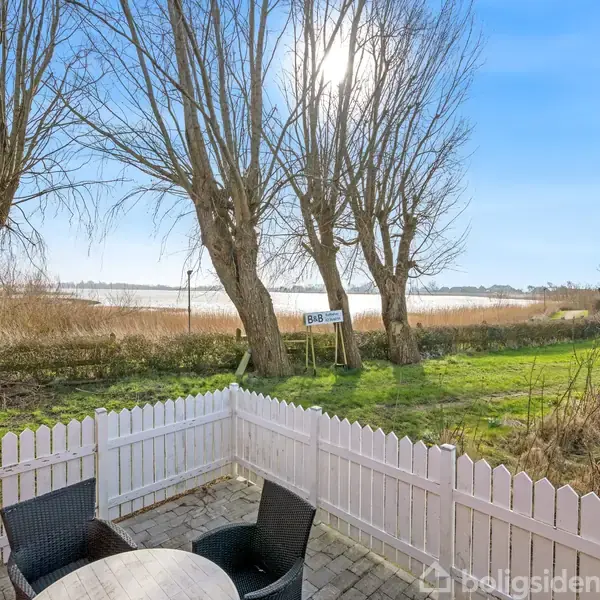 En terrasse med et rundt bord og sorte kurvemøbler omgivet af et hvidt stakit, udsigt til en sø gennem nogle træer i et landskabeligt område.