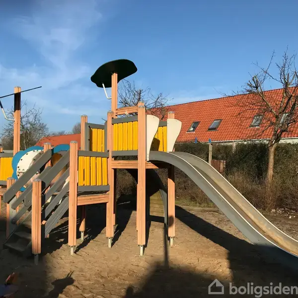 Legepladsstruktur med klatrestige og rutsjebane i et solrigt område med omgivende træer og et hus med rødt tag i baggrunden.