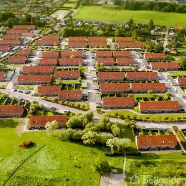 Rækkehuse med røde tage står organiseret i parallelle rækker. Husene har tilstødende haver, og området er omgivet af grønne marker og træer.