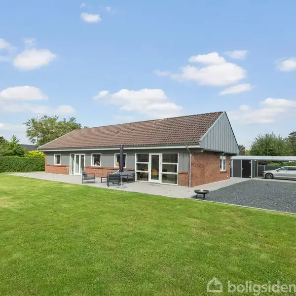 Et enkelt enfamiliehus med rødt tegltag og terrasse møbleret med sofa og bord, står i en velholdt have. En gyngestativ ses til venstre, bilparkering til højre.