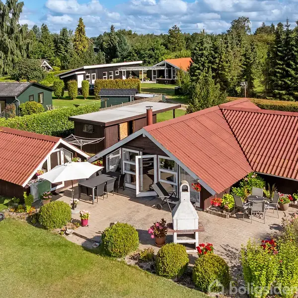 Et parcelhus med røde tegltage står i en velplejet have med terrasser og havemøbler. Omgivet af grønne buske og træer er der flere tilstødende bygninger og blåtskinnende himmel.