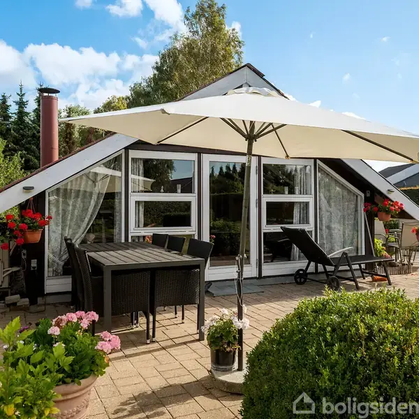 Hus med store vinduer; veranda med parasol, bord, stole og blomsterkrukker; optrukket gardiner. I baggrunden er træer og blå himmel.