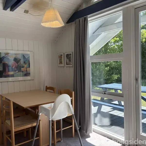 Et spisebord med stole står i et lyst rum med træpaneler på væggene. Over bordet hænger en lampe, og der er kunst på væggen. Store vinduer viser en have med picnicbord udenfor. Ingen tekst fundet.