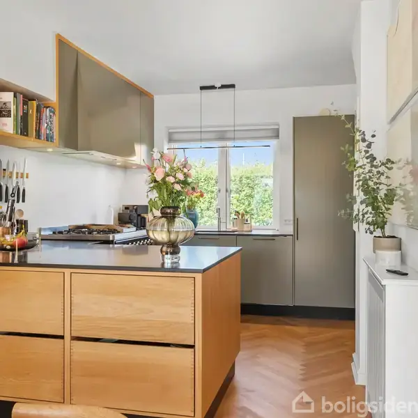 Køkkenø med træskuffer, blomster i vase ovenpå, omgivet af knive på holder. Bagved er der et vindue med udsigt til grønne buske og en lukket grå dør.