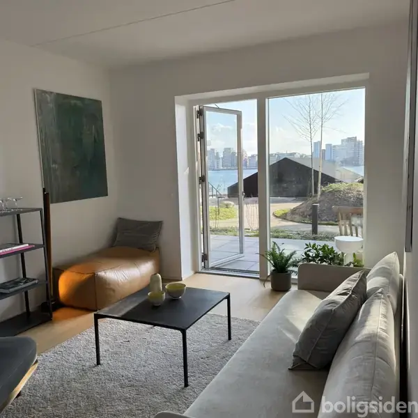 En lys stue med en grå sofa og et sort sofabord, der står foran en åben glasdør. Udenfor ses en udsigt over bygninger og natur.