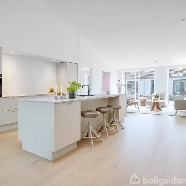 Køkkenø med barstole står i et lyst, moderne køkken og opholdsrum. Rummet har lyst trægulv, store vinduer og grønne planter. En sofa og et sofabord ses i baggrunden.