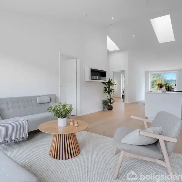 Stue med moderne, lys indretning. Grå sofa og lænestol, rundt træbord med planter. Baggrund viser et køkken med ø-køkken og store vinduer, der giver udsigt til haven.