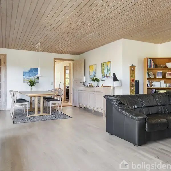Sort lædersofa står i hjørnet af en lys stue ved siden af et sofabord. Trægulv og loft giver varme, mens en spiseplads med træbord og stole er synlig i baggrunden.