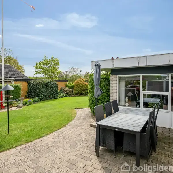 En terrasse med et sort spisebord og seks stole står stille. Baggrunden viser en grøn have med græsplæne, planter og en flagstang. En hvid bygning er delvist synlig.