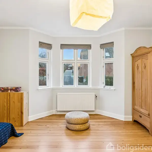 Et værelse med en enkelt seng til venstre og et træskab til højre, begge placeret på et lyst trægulv. Et rundt tæppe ligger foran et buet vindue med hvid radiator under.