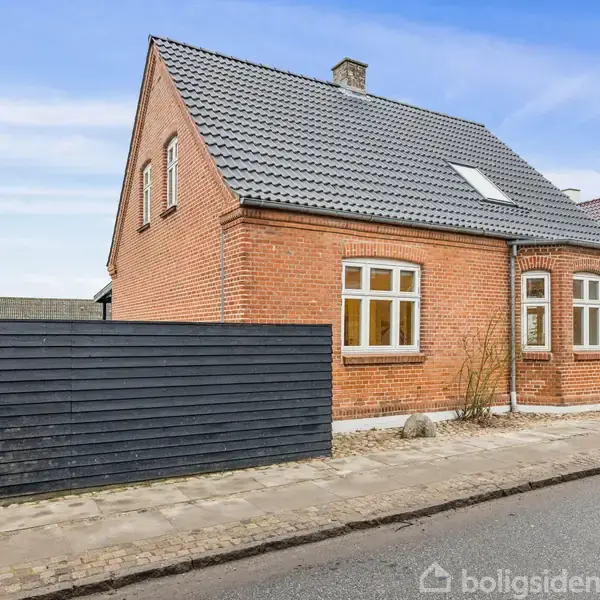 En rødstensvilla med sort tegltag står ved en fortov langs en vej. Huset har flere vinduer og en sort træplankehegn mod vejen. Himlen er blå.