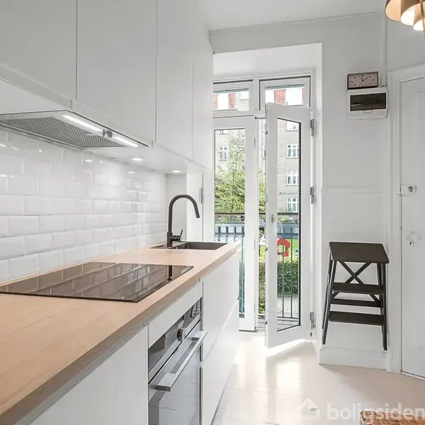 Et køkken med træbordplade og kogeplade. Døren til en balkon står åben. En stol står ved døren. Hvide fliser på væggen.
