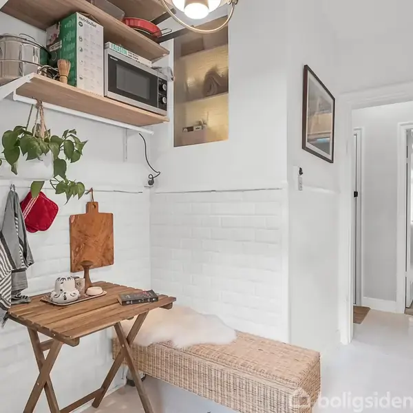 Et lille køkkenområde med et sammenklappeligt træbord fungerer som spiseplads. Der er en hylde med en mikrobølgeovn og planter ovenfor. En bænk med hynde står ved siden af. I baggrunden ses en gang.