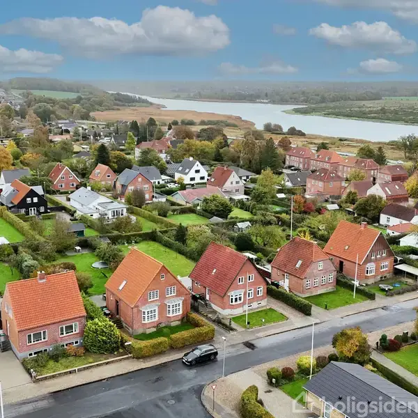 Rødtagede huse står på række langs en vej i et boligområde med velholdte haver. I baggrunden skinner en flod gennem det grønne landskab under en blå himmel med skyer.