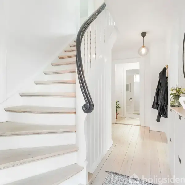 Trappe med sort gelænder fører opad i en lys entré. Omgivelserne inkluderer et spejl, en kommode med planter og jakker hængende på væggen. Lyse trægulve dækker rummet.