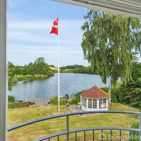 Et åbent vindue med udsigt til en sø og flagstænger med danske flag. En pavillon med rødt tag ses til højre, omgivet af græsarealer og træer.