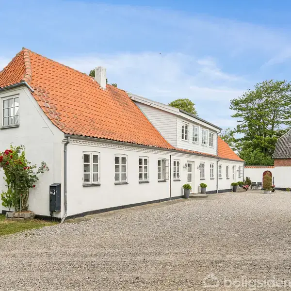 Hvid, to-etagers bygning med rødt tegltag står i en gårdsplads dækket af grus. En hæk og blomster til venstre ved siden af en træstruktur. Klar blå himmel over hovedet.