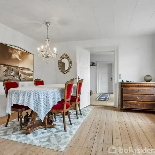 Spisebord med hvid dug og røde stole står under en lysekrone. Til venstre hænger et billede. Til højre en kommode og et vindue med lette gardiner.