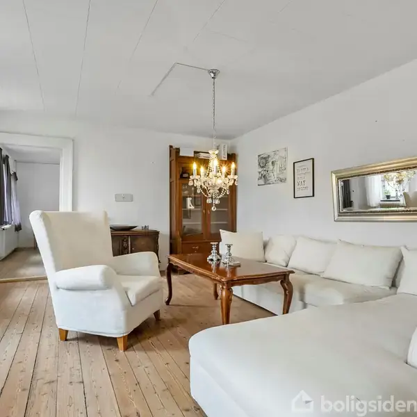 En lys stue med en hvid hjørnesofa og en lænestol placeret på et trægulv. Et træbord står foran sofaen, og en krystallysekrone hænger fra loftet. En stor spejlramme pryder væggen. Gardiner hænger ved vinduerne.