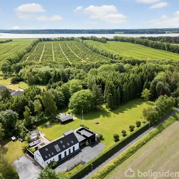 Et hvidt hus med sort tag står omgivet af frodig have og skov. I baggrunden ses store landbrugsmarker og en fjord under en klar, blå himmel.