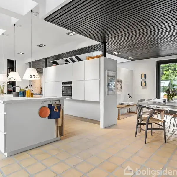 Køkken med hvide skabe og en central ø, lys fra hængelamper. I baggrunden er der en spiseplads med et bord og stole, omgivet af store vinduer, der viser en grøn have.