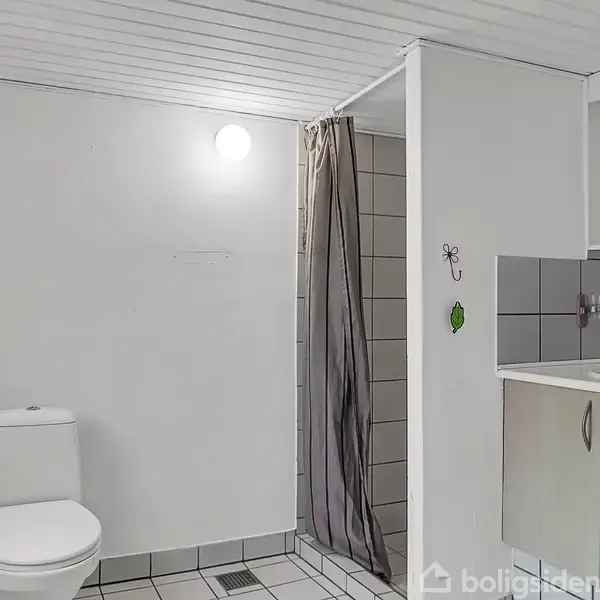 Toilet placeret ved siden af en lille duschkabine med grå fliser; vaskearrangement med opbevaring til højre, lyse vægge og loft i et funktionelt badeværelse.