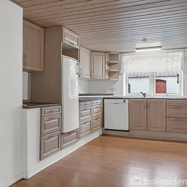 Køkken med træskabe og hvidevarer, herunder køleskab og opvaskemaskine, i en lys og rummelig indretning. Store vinduer med udsigt til udendørs område. Trægulv og træloft.