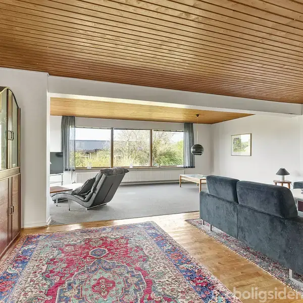 En stue med sort sofa og stol, placeret på et farverigt orientalsk tæppe. Store vinduer lukker lys ind, omgivet af hvide vægge og træloft, med reol i baggrunden.