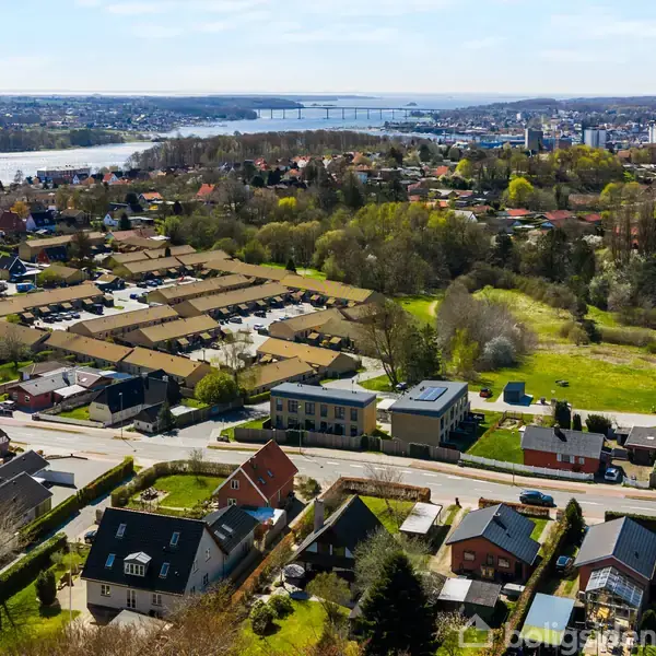Byområde med huse strækker sig mod en flod i baggrunden, hvor en bro forbinder bydele. Landskabet er blandet med beboelse og grønne områder. Klart himmeloverblik.