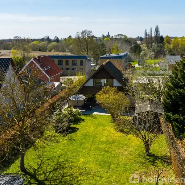 Et hus med en markant spidst tag ses i en velholdt have. Forhaven har en græsplæne og træer. Omkringliggende huse og åbne områder udfylder baggrunden.