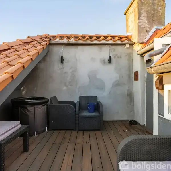 En tagterrasse med trægulv, hvor der står to grå loungesofaer og et rundt sort bord. Omkranset af orange tegltage og en hvid murvæg.