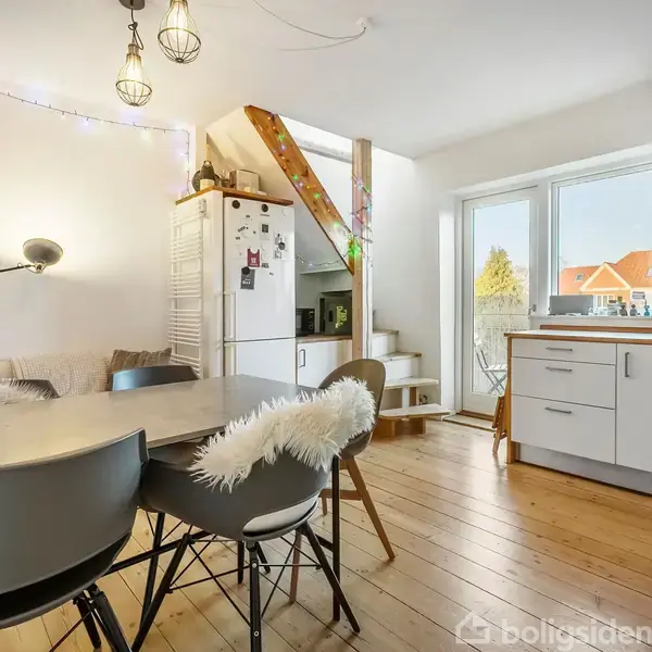 Et moderne køkken-alrum med spisebord og stole foran. Køkkenet har hvide skabe, trægulve og en trappe til venstre for køleskabet. Lysekrone og lyskæde oplyser rummet.
