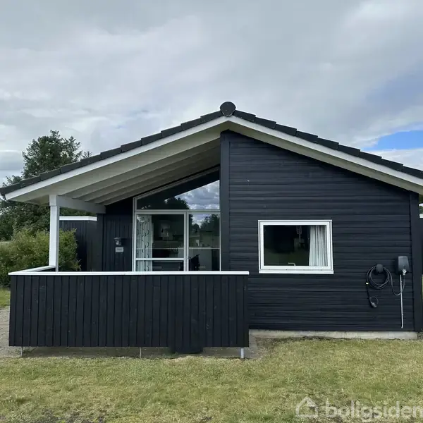 Et sort træhus med veranda står på en græsplæne, omgivet af buske og træer under en overskyet himmel. En enkelt hvid ramme om vindue bryder det mørke ydre.