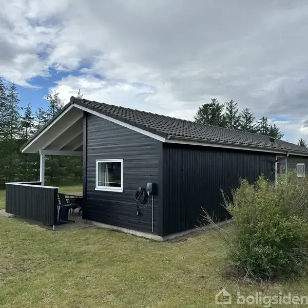 Sort træhus med hvide vinduesrammer står på en græsplæne i nærheden af træer. Terrasse med bord og stole ved siden af huset. Skyet himmel ovenfor.