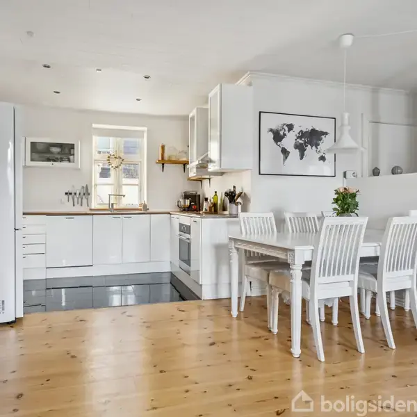 Et hvidt, moderne køkken med integreret spiseplads. Bordet har seks hvide stole. Vægdekoration inkluderer et verdenskort. Trægulv og loftslamper i et lyst rum.