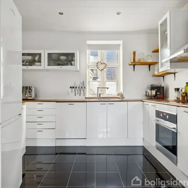 Et lyst og moderne køkken med hvide skabe og sorte gulvfliser, indeholder en ovn, køleskab og mikrobølgeovn. Et lille vindue i midten tillader naturligt lys at komme ind.