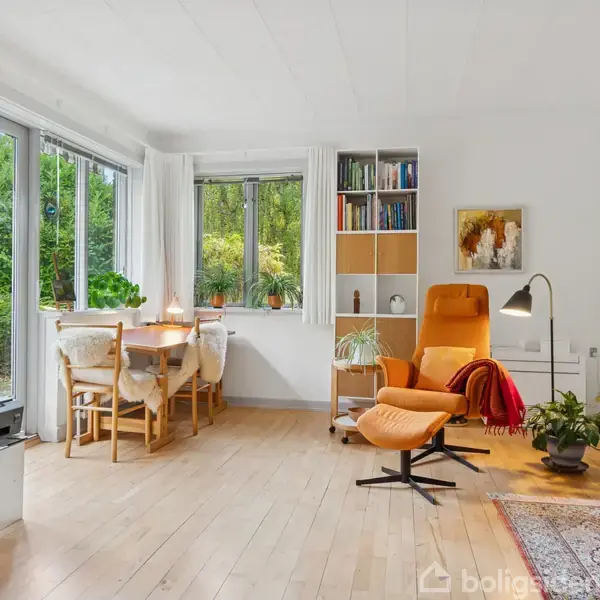 Stue med lænestol i orange ved siden af gulvlampe, reol med bøger og planter. Lyst rum med store vinduer, spisebord med stole og udsigt til have.
