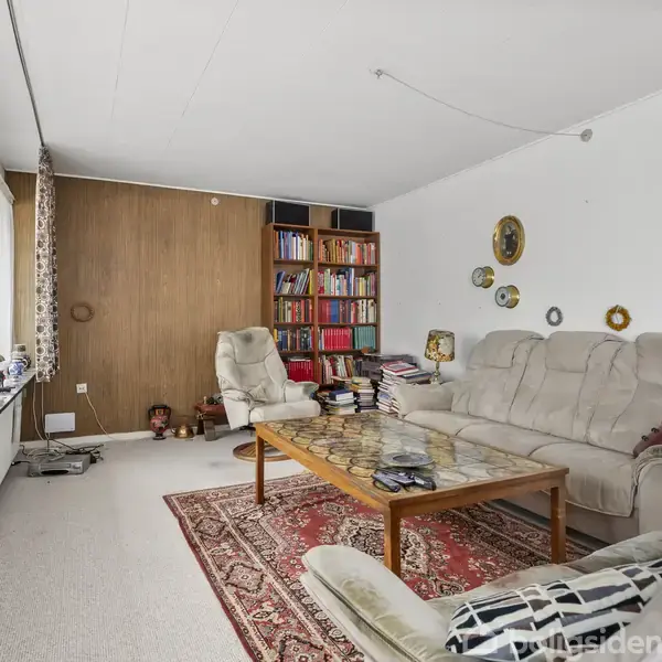 Stor stue med beige sofaer omkring et træbord. Rød tæppe dekorerer gulvet. Farverige bøger fylder en reol i hjørnet. Væggen har ovale dekorationer, og gardiner hænger ved store vinduer.
