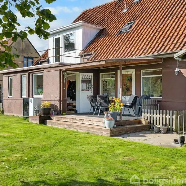Et rødt teglhustag på et hus med terrasse. To stole og blomsterkrukker står på terrassen. Græsplæne og træer i forgrunden.