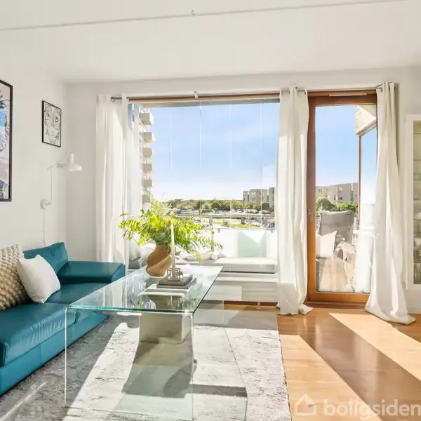 Blå sofa med puder står ved siden af et glasbord med en plante. Store vinduer viser en balkon med udsigt. Væggen har en plakat med teksten "PRECIOUS LIBERTY".
